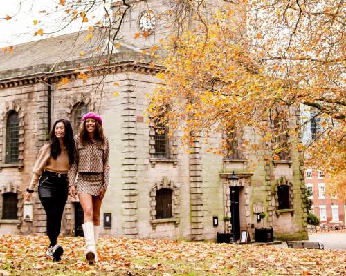 Two friends walking through a park in autumn with fallen orange and red leaves with St Phillip's Cathedral in the background