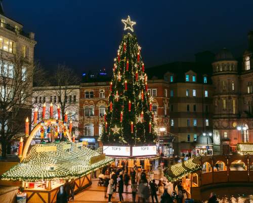 A tall Christmas tree stands in a square in Birmingham at night, the lights are lit and people are gathered round its base at Christmas market stalls