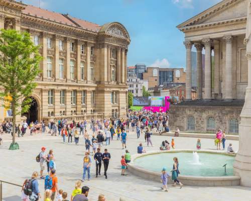 Groups of people sitting outside in summer clothes. Surrounded by Chamberlain Square fountain and Birmingham Museum and Art Gallery