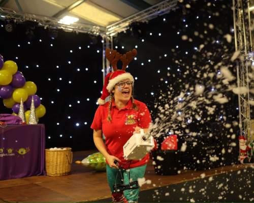 A worker at the big sheep wearing a christmas hat and antlers spraying fake snow at a christmas party