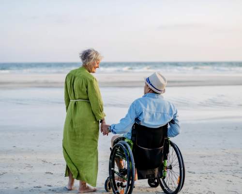 An elderly couple holds hands on a sandy beach. The woman in a green dress walks beside a man in a wheelchair, both gazing at the calm sea.