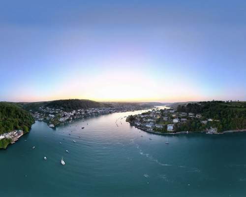 Aerial view of a tranquil river winding between two lush, green hills, dotted with small boats. Houses line the riverbanks under a clear, pastel sunset sky.