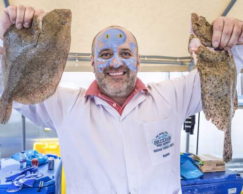 Smiling man with face paint holds a large fish in each hand, wearing a white coat with a market logo. Bright, cheerful market setting.