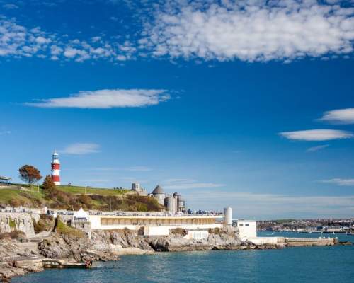 A coastal scene with a red and white lighthouse on a grassy hill, blue sky with scattered clouds, rocky shore, and calm blue sea creating a serene mood.