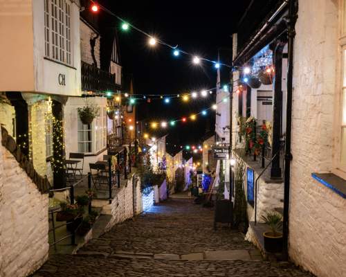 A charming cobblestone street at night, lined with cosy houses adorned with glowing multicoloured string lights, creating a festive, warm atmosphere.