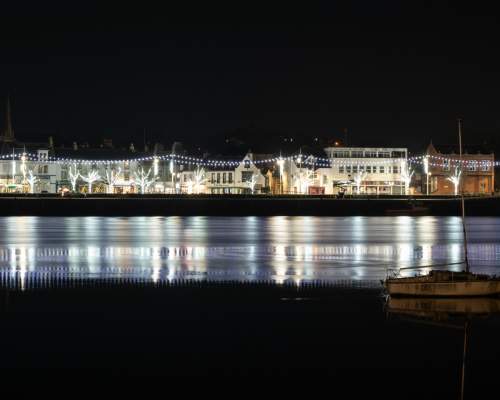 Nighttime waterfront scene in Bideford, North Devon, with buildings adorned with festive lights and their reflections on the water. A lone sailboat is silhouetted in the foreground, creating a serene atmosphere.