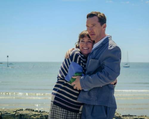 A couple embraces warmly by the beach under a bright blue sky. Behind them, the calm sea and a distant sailboat set a peaceful, joyful tone.
