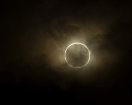An annular solar eclipse hangs in a dim, hazy sky, with the Moon covering the centre of the Sun and leaving a bright, glowing ring of light around its silhouette. The surrounding clouds appear muted and softly illuminated by the eclipse’s orange halo.
