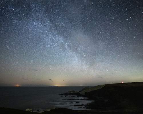 A star‑filled night sky over the Hartland coastline in North Devon, with the Milky Way stretching diagonally above dark cliffs and a calm sea. Faint lights sit low on the horizon, contrasting with the rugged shoreline and the bright band of stars overhead.