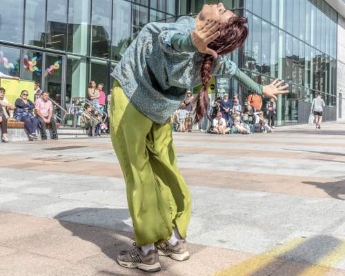 A street dancer in a teal top and lime-green trousers arches back with arms out, performing before spectators outside the Box Plymouth building. The scene feels lively.