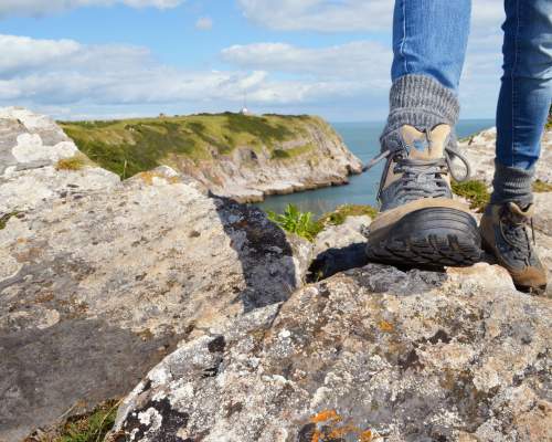 A person stands on a rocky cliff, wearing hiking boots and jeans. A scenic view of rugged terrain and blue sea stretches under a partly cloudy sky.