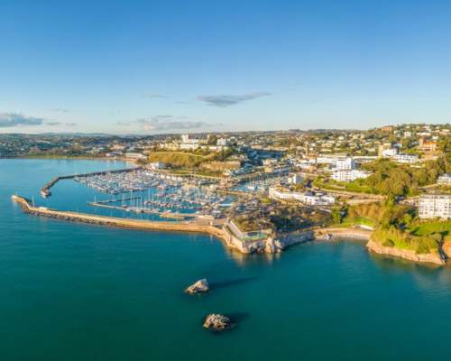 Aerial view of a coastal town with a marina full of boats, surrounded by lush greenery and buildings. The calm blue sea and clear sky create a serene atmosphere.