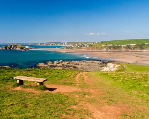A wooden bench overlooks a coastal landscape with turquoise waters, sandy beaches, and distant green hills. The scene conveys tranquillity and natural beauty.