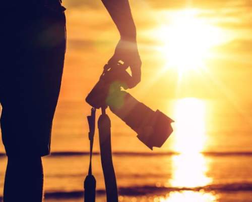 image shows person stood on the beach with camera in front of sunset