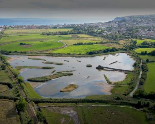 Aerial view of a scenic wetland area with ponds, surrounded by vibrant green fields and distant hills. A tranquil landscape under a clear sky.