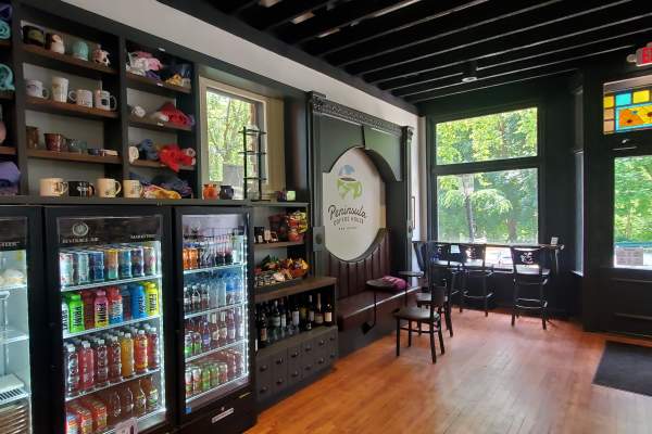 Peninsula Coffee House interior showcasing shelves with mugs, a fridge stocked with drinks, and large windows overlooking greenery.