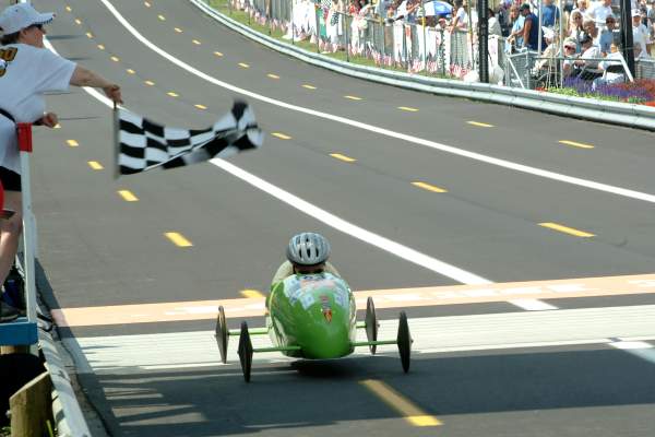 A person in a green soapbox derby car crosses the finish line, as an official waves a checkered flag in an Akron race with spectators watching.