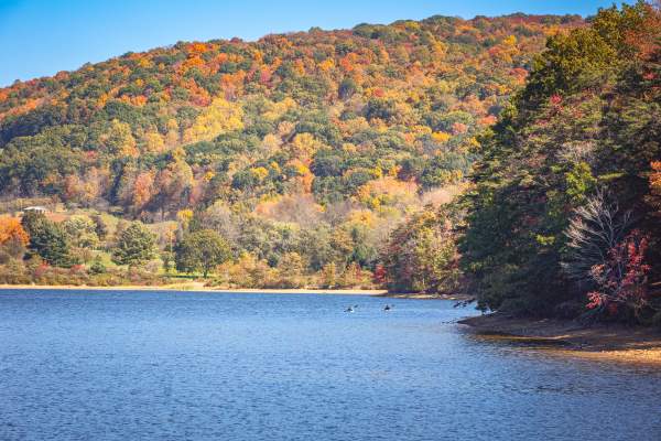 A mountain of trees shining in reds, oranges, and golds sits as the backdrop to the blue waters of Lake Habeeb. Two people are seen off in the distance in a canoe.