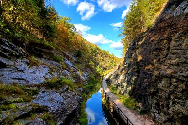 A scenic canal towpath runs alongside still water with dramatic rock walls and bright blue sky overhead.