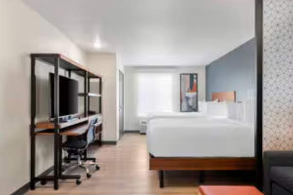 A modern hotel room featuring two queen beds with white linens, a work desk with a large TV, and a cozy sitting area separated by a patterned privacy screen. Light wood floors and clean lines give the space a bright, contemporary feel.