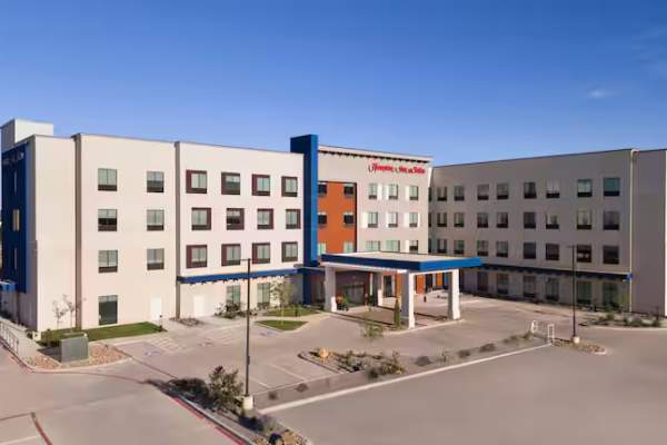 A modern Hampton Inn & Suites hotel with a clean, contemporary exterior featuring a mix of white, beige, and navy blue tones. The four-story building has large windows, a covered entrance, and a well-maintained parking lot with neatly landscaped areas. The clear blue sky in the background highlights the hotel's inviting and spacious design.