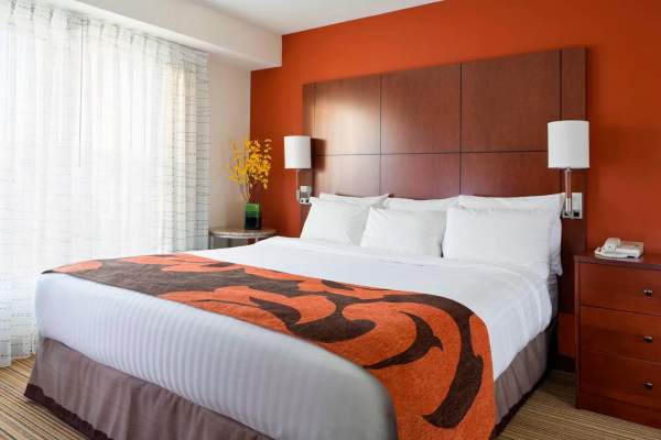a bright, modern hotel room in Amarillo with a king bed, crisp white linens, and a bold orange accent wall, featuring a patterned runner, bedside lamps, and soft natural light filtering through sheer curtains
