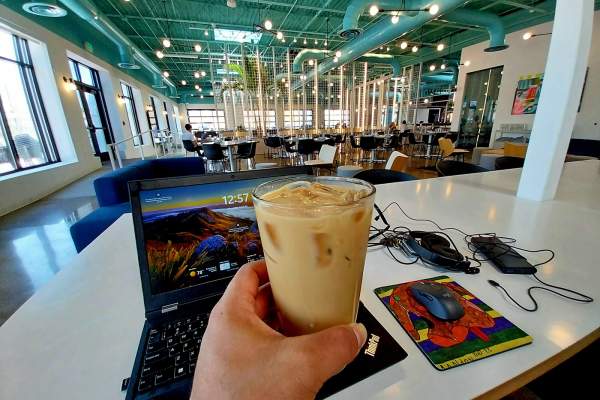 Person holds coffee from first-person point of view. Behind the coffee, a laptop and book sit on a table in a coworking space.