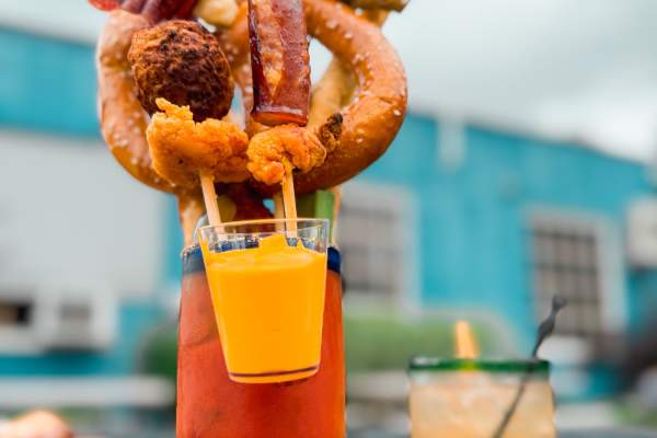 two drinks sit on a table outside. One drink has a pretzel, salami, and edible foods sticking out fo the drink.