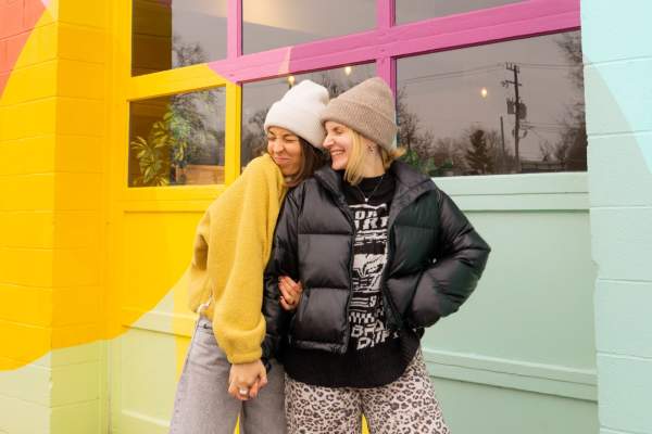 Two women wearing beanies and coats hold hands and stand in front of a colorful mural.