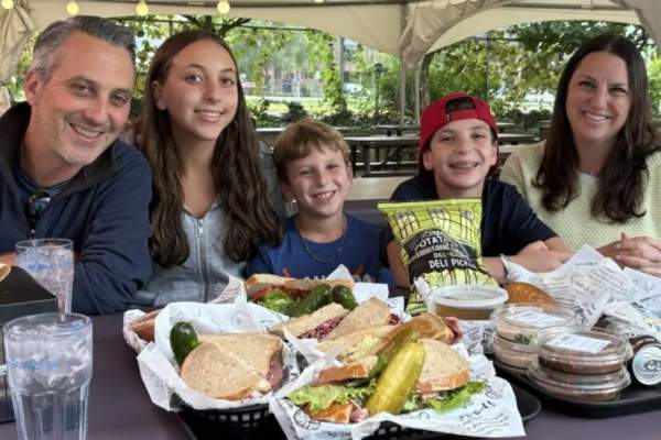 Family of five eat outdoors at a table full of sandwiches and sides.