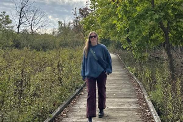 Woman walks outdoor on wooded boardwalk passing over a greet meadow with trees in the background.