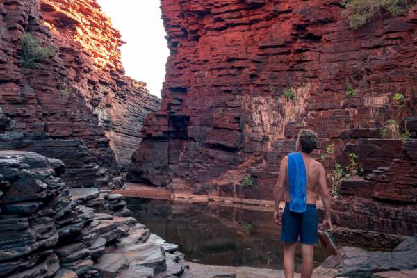 A man walking through Joffre Gorge in Karijini. There is a pool in front of him and you can see the sky through a gap in the gorge walls.