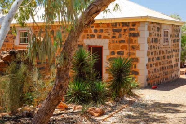 Three people walking alongside a restored bluestone building in Cossack, Western Australia, with water visible in the background.
