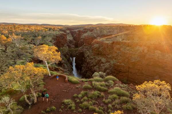 A family walks towards the Joffre Gorge Lookout in Karijini National Park - a waterfall is visible from the lookout and it's surrounded by red rocks, spinifex and snappy gum trees.