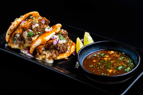 Birria dipping sauce in a bowl plated with tacos featuring a bun-like shell, stuffed to the brim with meat, onions, and cilantro.