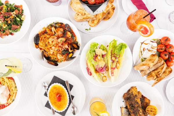 Lucca Coastal Kitchen variety of food offerings on plates with drinks in between on a plain white table.