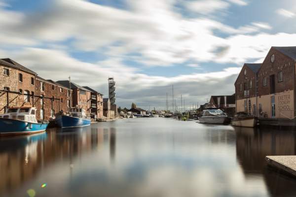 A serene waterfront scene with moored boats on a calm river, flanked by rustic brick buildings under a partly cloudy sky. A peaceful, charming atmosphere.