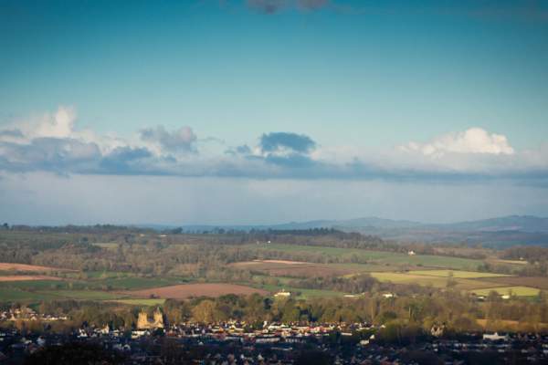 A picturesque rural landscape with patchwork green fields, distant rolling hills, and a small town in the foreground under a vast blue sky with scattered clouds.