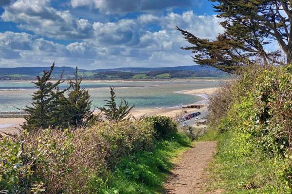 A scenic coastal path leads to a sandy beach, bordered by lush greenery and trees. The sea sparkles under a partly cloudy sky, conveying a serene atmosphere.