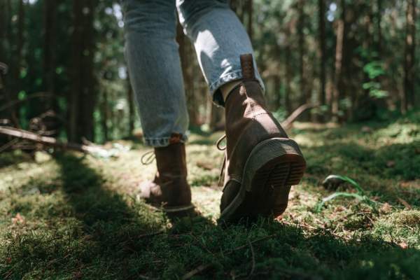 Close-up of a person wearing brown boots and blue jeans walking on a sunlit, moss-covered forest floor, conveying a sense of adventure and tranquility.