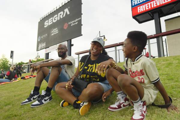 Family sitting on a grassy hill at Segra Stadium in Fayetteville, smiling and talking during a baseball game