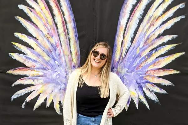 A smiling woman in sunglasses poses in front of a colorful painted mural of angel wings on a black backdrop in Fayetteville, NC. The wings are lavender with accents of yellow, pink, and white, creating a vibrant, photo-worthy moment.
