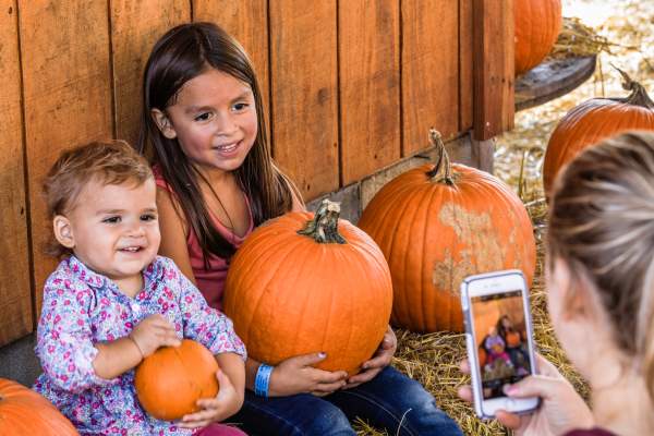 Two young children sit among pumpkins smiling while an adult takes their photo on a smartphone, capturing a festive fall selfie moment in Fayetteville.