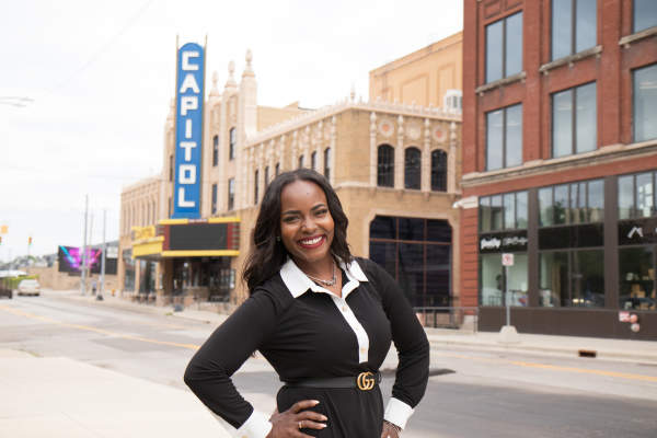 A woman dressed in a black and white long-sleeve shirt with a belt around the waist stands in front of a historic theatre.