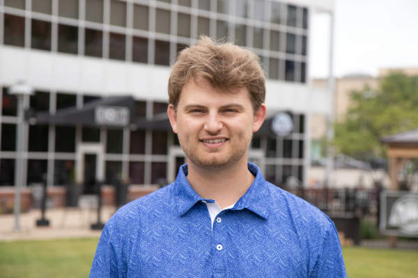 A young man wearing a blue golf shirt stand in front of a restaurant that is part of a multi-use building