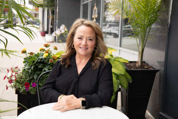A woman with long blonde hair wearing a long-sleeve black blouse sits at an table outside a restaurant. Large containers of summer-blooming flowers are behind her.