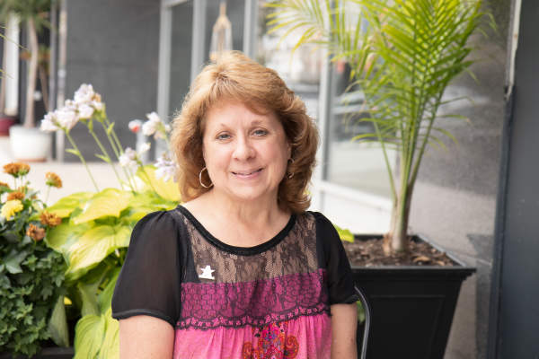 A woman with mid-length auburn hair wearing a short-sleeve pink top with black lace sits at an outside restaurant table with containers of large hostas and flowers behind her.