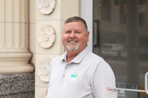 A man with a salt & pepper hair and beard wearing a light grey golf shirt stands in front of a historic building with marble columns.