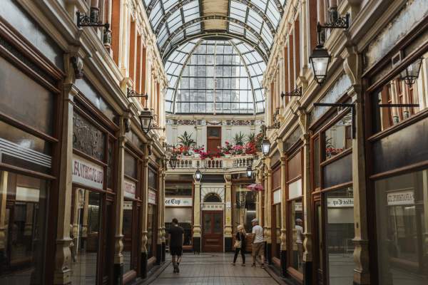 Hepworth Arcade in the summer sunshine. The arched windows show the tower of Hull Minster in the background. Below, the lovingly tendered plants and flowers sit on the windowsill. Shoppers stroll the shops and Victorian lamps line the doorways.