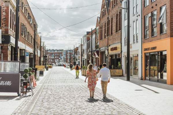 Humber Street on a sunny day facing out to the marina. The brightly coloured shop fronts lead into the distance where you can just see boats bobbing on the water. Lights are strung across the road from roof to roof. A couple strolls away from the camera, the lady wears a summer dress and the man is in wearing light chinos and a white shirt.